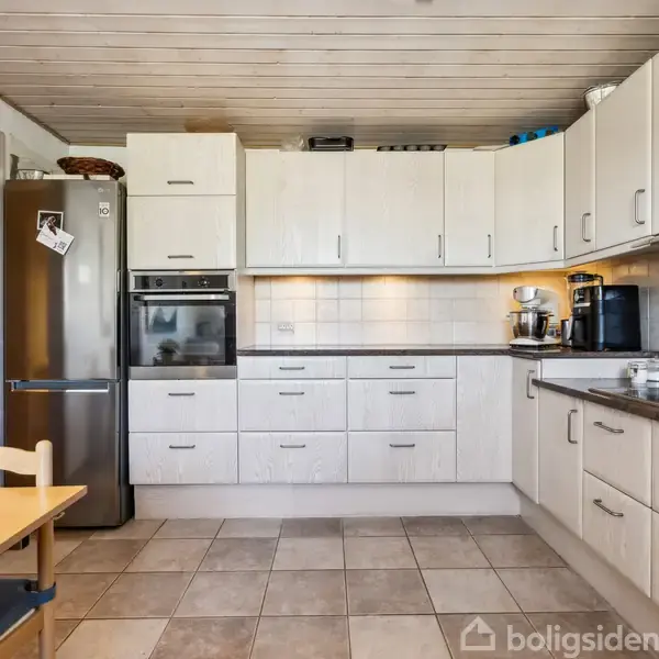 Køkken med vask og komfur på køkkenbordet, hvide skabe og skuffer omkranser rummet. Spisebord til venstre og stor køleskab ved siden af ovn i hjørnet.