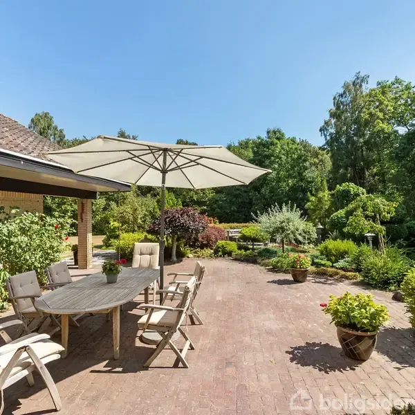 En terrasse med træmøbler og parasol foran et hus. Stolene står omkring et ovalt bord. Haven omkring har tætte, grønne buske og træer under en klar blå himmel.