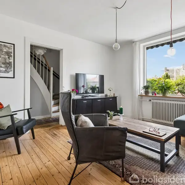 Stue med lænestol og sofa omkring sofabord på tæppe. TV ovenpå skænk foran stor vindue med udsigt til have. Trappe i baggrund ved åben dør. Ingen synlig tekst.