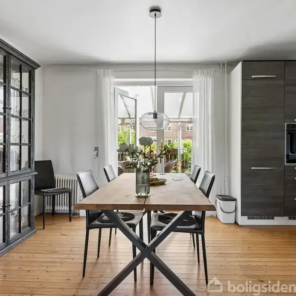 Et spisebord med blomster i en vase står i en spisestue. Rummet har et højt skab til venstre, moderne stole omkring bordet, og udsigt til have gennem en åben dør.