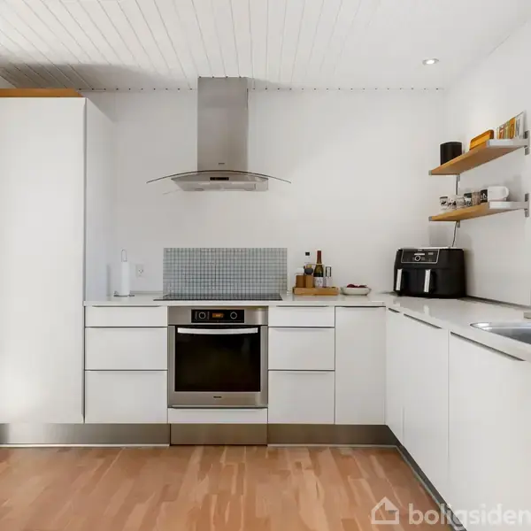 Et moderne køkken med hvide skabe, en indbygget ovn, emhætte, og åbne hylder. Der står en airfryer og nogle krydderier på bordet. Trægulv og vindue til højre.