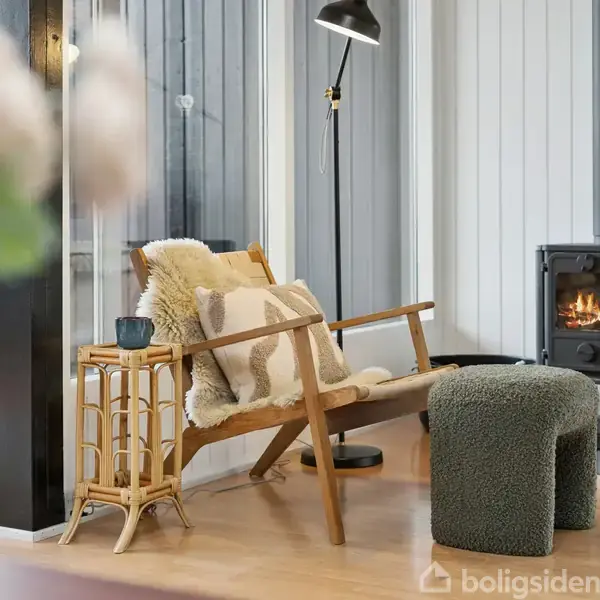 En komfortabel lænestol med tæppe og pude står i et hyggeligt rum ved siden af en lille, sort brændeovn. Et tæppe og bordlampe supplerer den varme, moderne indretning.