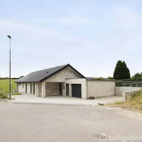 Et moderne hus med garage ses fra vejen, omgivet af åbne marker og en træbevokset baggrund. Det er en lys murstensbygning med et mørkt tag under en klar himmel.
