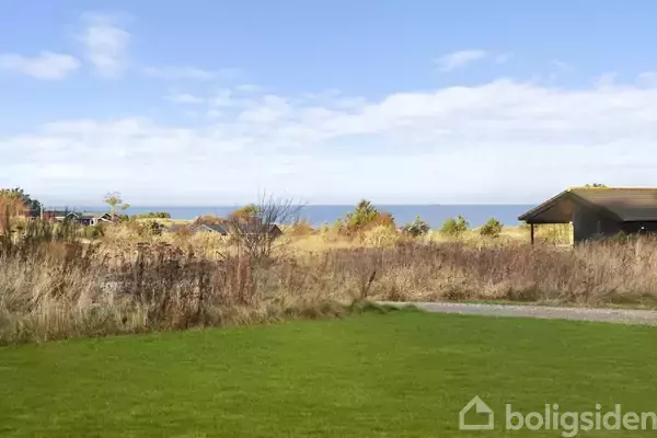 Grøn græsplæne strækker sig mod kysten, hvor havet møder himlen i horisonten. Omgivet af buske og en træhytte til højre.
