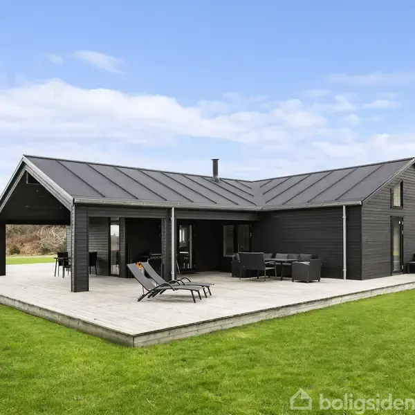 Et moderne træhus med sort facade står på en træterrasse. To liggestole og et sæt havemøbler er placeret foran huset. Græsplæne omgiver huset.