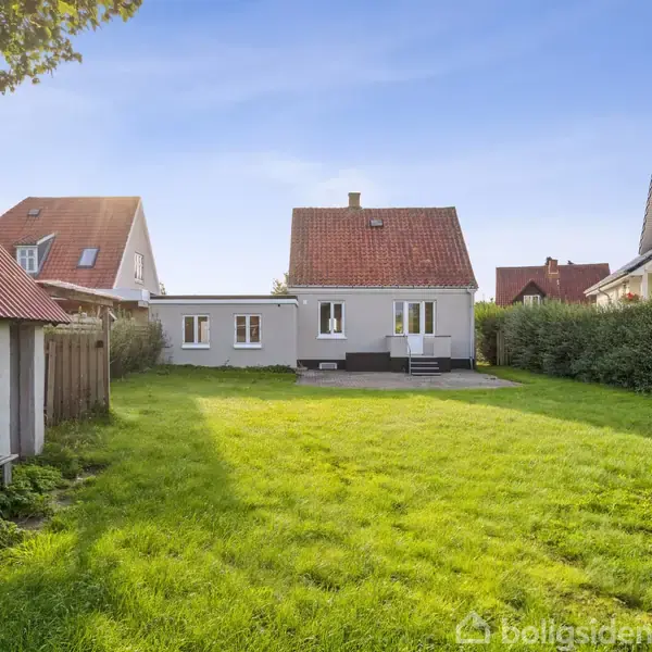 Et hvidt hus med rødt tag står i en baghave med velplejet græsplæne omkranset af hække. På højre side ses en terrasse med trapper. Sollys skinner på området.