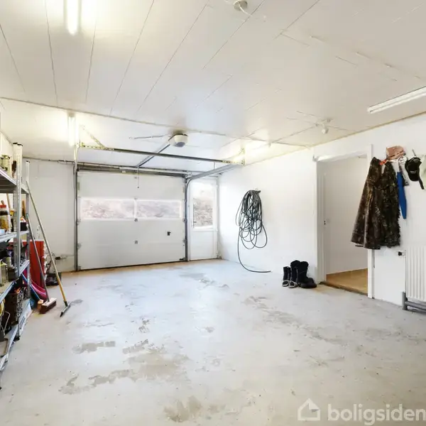 En stor garage med hvidmalede vægge og et betonlignende gulv. Til venstre er der metalreoler med værktøj og materialer. En åbnet garageport ses for enden, og jakkekroge hænger til højre.