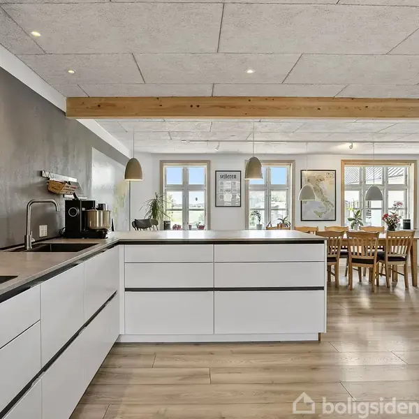 Moderne køkken med hvide skabe, centralt køkkenbord og indbygget komfur. I baggrunden ses en spisestue med træbord og stole, store vinduer, flere planter og ophængte billeder på væggen.