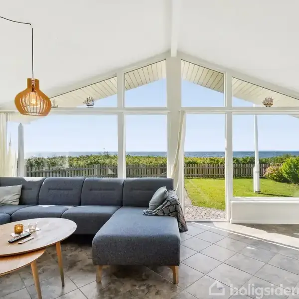 En grå hjørnesofa står foran store vinduer med udsigt til en grøn have og havet. En rund trælampe hænger over et sofabord med en vase.