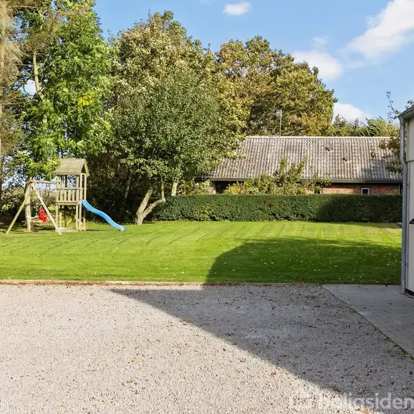 En legeplads med en rutsjebane står på en velplejet græsplæne foran et træhus. Området er omgivet af træer under en klar blå himmel.