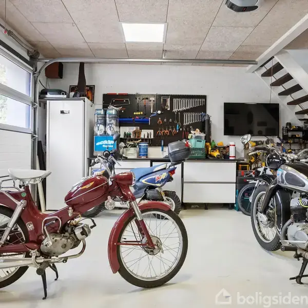 Røde og sorte motorcykler står stille i en velordnet garage. Et værkstedsområde med værktøj og reservedele ses i baggrunden, mens en trappe fører op til en anden etage.