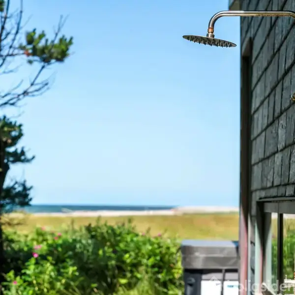 Udedusch fastgjort til en husmur, vender mod en frodig have med træer. I baggrunden ses en strand og blå himmel, hvilket skaber en afslappet atmosfære.