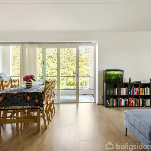 En stue med spisebord og stole med blomster på, en fisketank og bogreol. Rummet har store vinduer, der fører ud til en balkon med grøn udsigt.