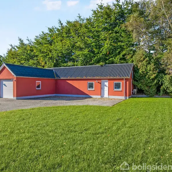 Rød bygning med grå tag ligger ved stor grøn græsplæne. Bygningen har flere vinduer og en garageport. Området er omgivet af høje træer og en klar blå himmel.