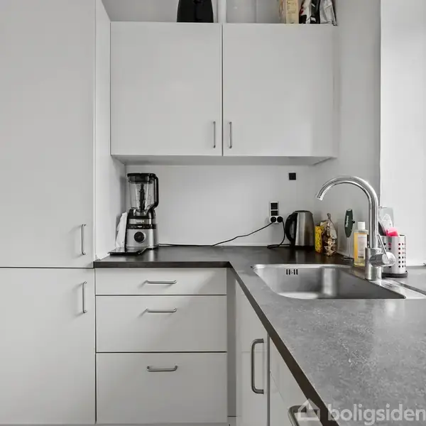 Et hvidt køkken med indbyggede skabe og skuffer, en bordplade i grå farve, en vask med en hane, og apparater som en kaffemaskine. Store vinduer giver naturligt lys.