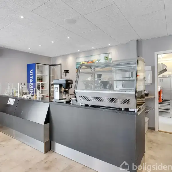 Sort disk med glasmontre, placeret i lyst rum med grå vægge. Kaffemaskine bag disken, åbent tilstødende køkken. Enkelt maleri på væggen og rød stol ved siden af.