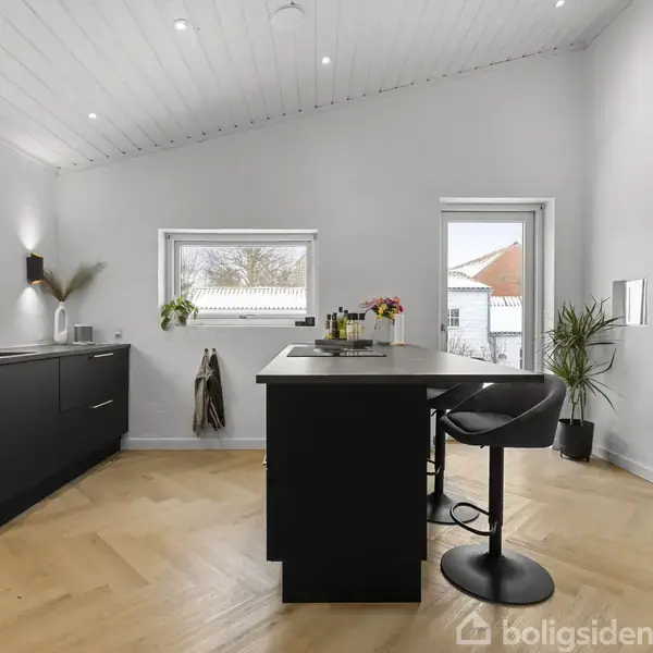 Køkken med sorte skabe står stille i et lyst rum. Midt i rummet er der en ø med høje stole. Udenfor vinduerne ses nogle huse og træer.