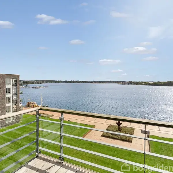 Altan med glasrækværk har udsigt over en rolig sø. Til venstre ses en moderne bygning med flere vinduer. Der er grønne områder og blå himmel med spredte skyer.