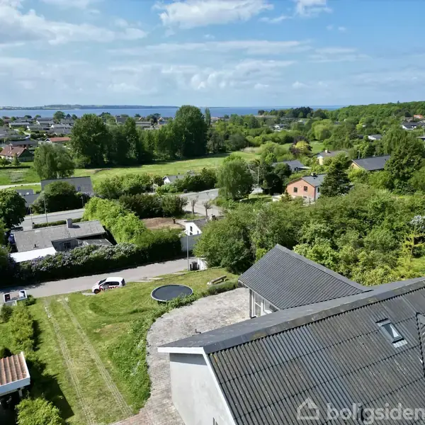 Hustag set ovenfra i et grønt beboelsesområde, rolig vej med biler, omkranset af træer og haver, og i baggrunden en udsigt til havet under en skyet himmel.