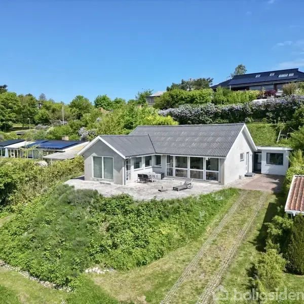 Et hvidt hus står på en græsplæne, omgivet af grønnende hække. Foran huset er en terrasse med havemøbler. Baggrunden indeholder flere huse og frodig bevoksning under en klar blå himmel.
