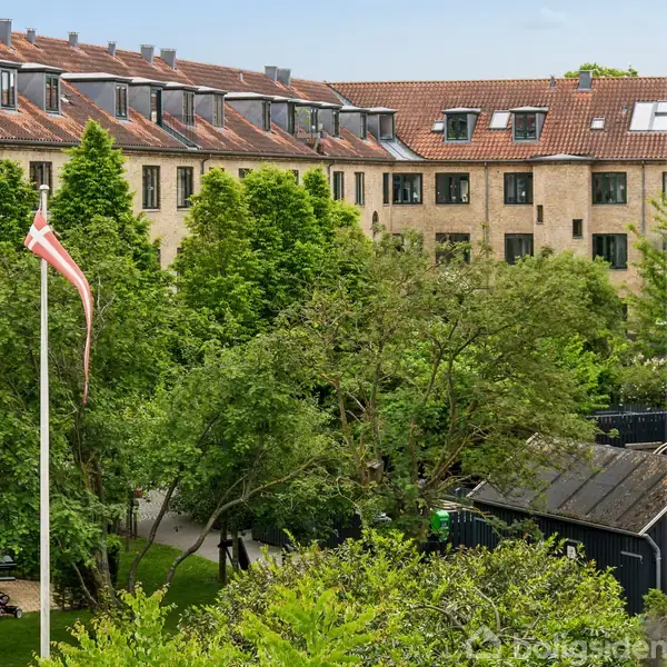 Bygning med rødt tegltag og mange vinduer står bag grønne træer i en gårdhave. Dannebrog på en flagstang vejrer i vinden. Blåt himmel ses i baggrunden.