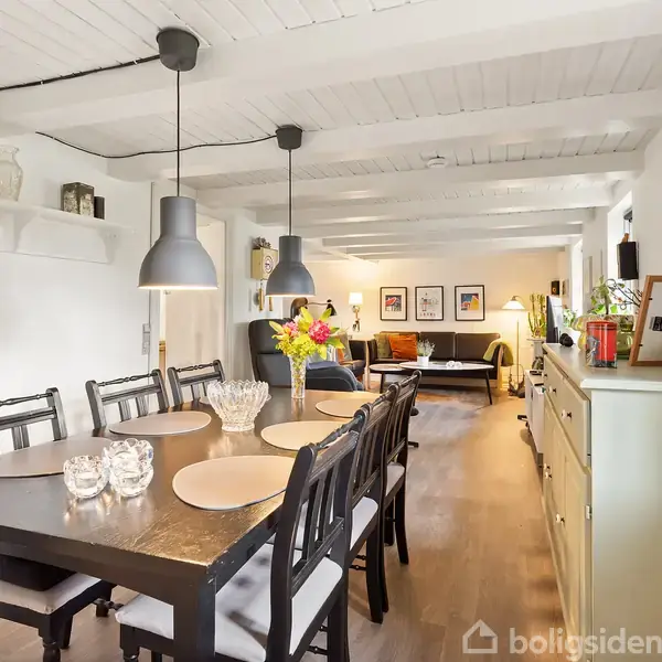 Spisebord med otte stole står i forgrunden, dækket med tallerkener og blomster. Over bordet hænger to pendellamper. I baggrunden ses en stue med sofaer og kunst på væggene.