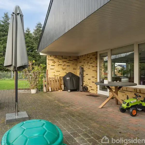 Terrasse med brune fliser og et bord. En grøn legetøjsbil og grøn oppakning ses. Omgivet af plæne og buske; store vinduer fører ind i huset.