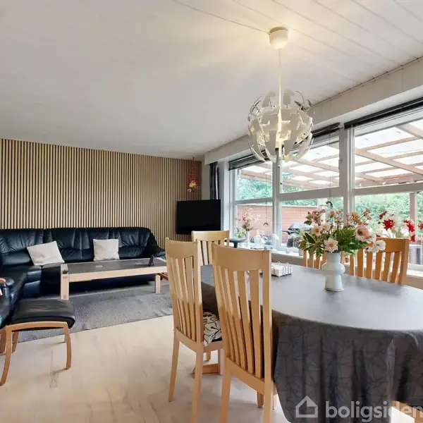 Stue med sort lædersofa arrangeret mod en væg af træpaneler. Spisebord med blomsterdekoration. Store vinduer viser en terrasse med grøn udsigt. Lampen hænger over bordet.