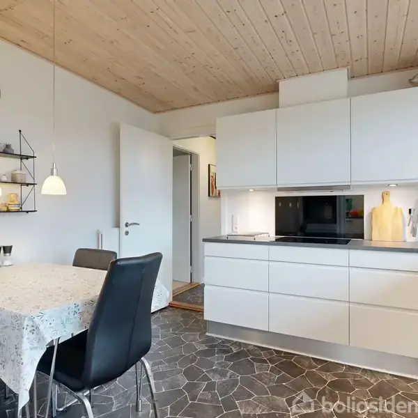 Et moderne køkken med hvide skabe og sort bordplade. Et spisebord med lys dug og sorte stole står ved siden af. En dør leder til en anden del af huset. En hylde med dekorationer hænger ved siden af en ur.