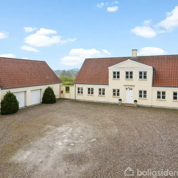 Et stort, hvidt, klassisk hus med rødt tegltag står på en grusplads. Til venstre er der en garagebygning i samme stil. Baggrunden består af åbent landskab under en blå himmel.
