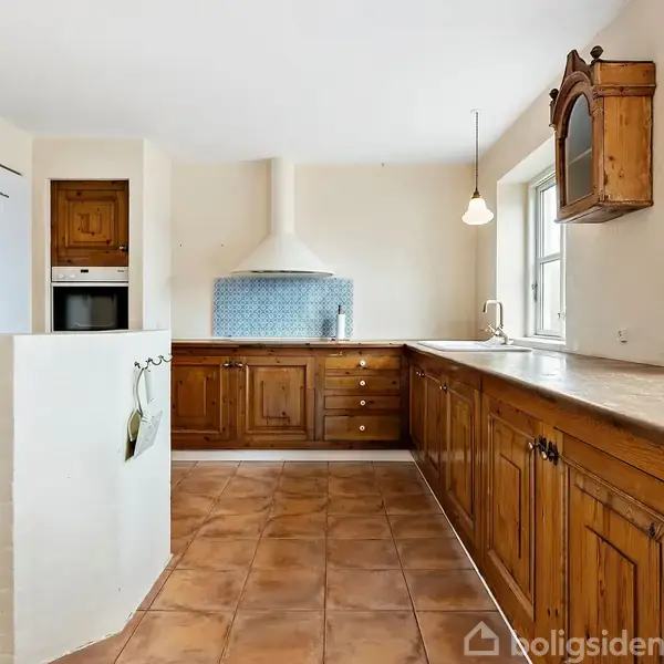 Et køkken med brune trækabinetter strækker sig langs væggene. Der hænger to lamper over en lys køkkenbænk. Et flisegulv og en indbygget ovn præger rummet.