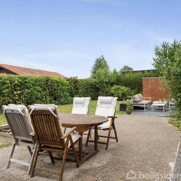 Træhavemøbler står på en fliseterrasse med borde og stole, omkranset af hække og grønne buske. En solskinsdag i en rolig have med en rød murstensvæg i baggrunden.