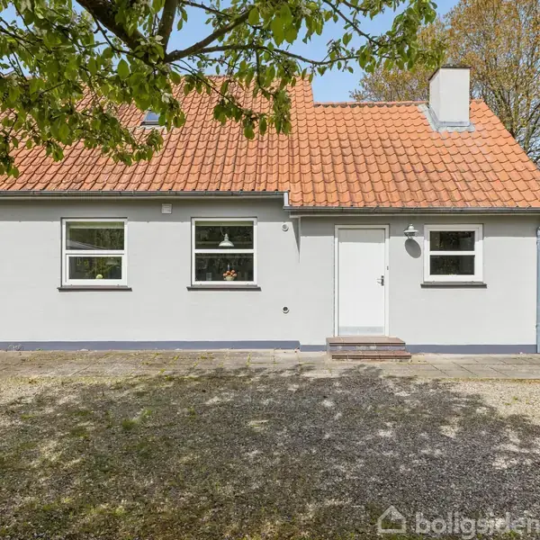 Et hvidt hus med rødt tegltag står omgivet af en grusindkørsel og frodig have. En garage ses i baggrunden under træer. Solen skinner klart i landskabet.