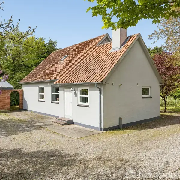Hvidt hus står ved en grusbelagt gårdsplads. I baggrunden ses en rød murstensgarage med en grøn buebusk foran. Omgivet af træer og blomstrende buske.