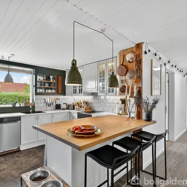 Et moderne køkken med en træbordplade og to sorte stole. Hængende lamper oplyser bordet. I baggrunden er der en opvaskemaskine, køleskab, vask og et stort vindue med udsigt til en have.