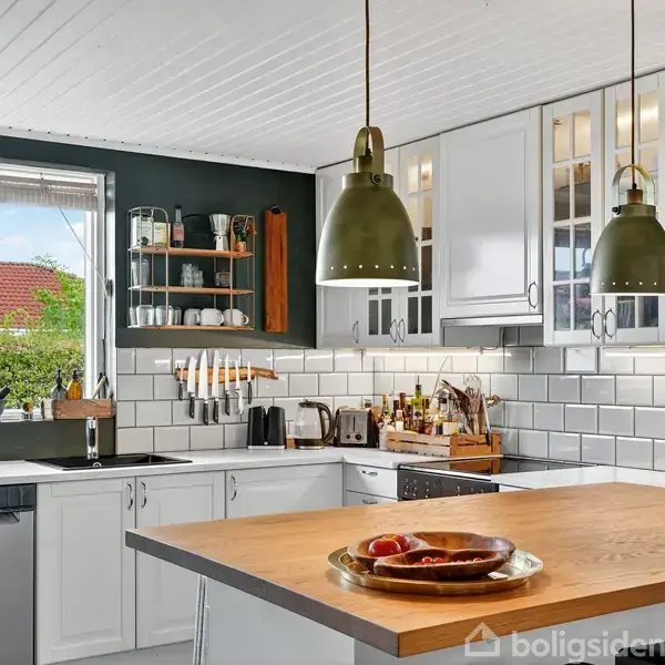 Et lyst køkken med hvide skabe og sort bordplade. En ø med træbordplade og pendellamper over. Kogegrej hænger på væggen. Et stort vindue viser en have udenfor.