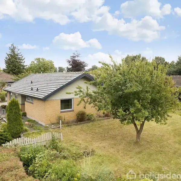 Hus i gule mursten med gråt tag, omgivet af en have med græs og træer. Beliggende i et villakvarter med flere lignende huse. Ingen tekst bortset fra "boligsiden" vandmærket.