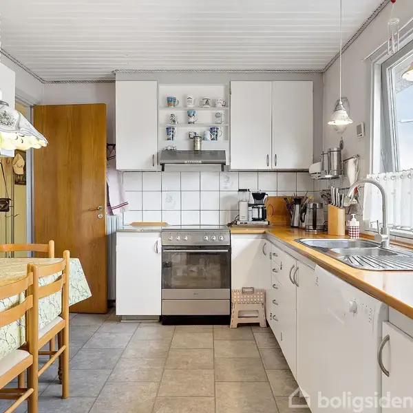 Køkken med hvide skabe og fliser. Komfur og emhætte er placeret centralt. Spisebord med to stole til venstre, og køkkenvask mod højre ved et bredt vindue.