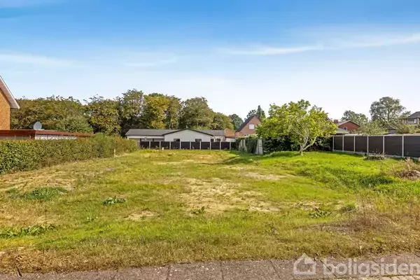 Græsplæne strækker sig med træer i baggrunden; omkranset af hække og huse. Klar blå himmel danner baggrund.