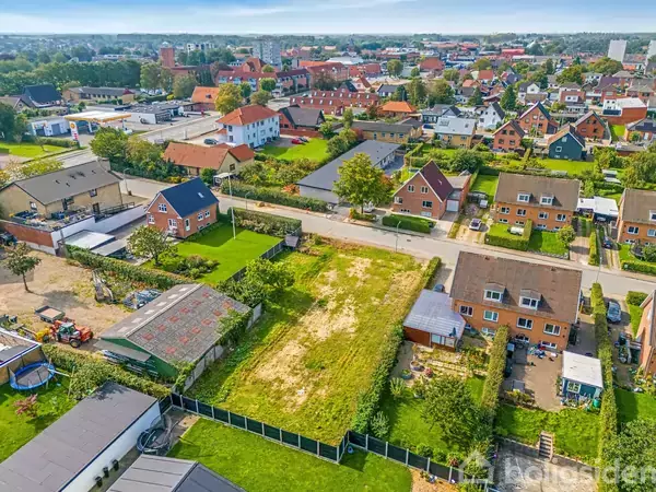 Tomt jordstykke ligger stille mellem huse i en fredelig forstad. Omkranset af små haver og boligblokke, strækker sig området i centrum af billedet med velholdte græsplaner og stiforbindelser.