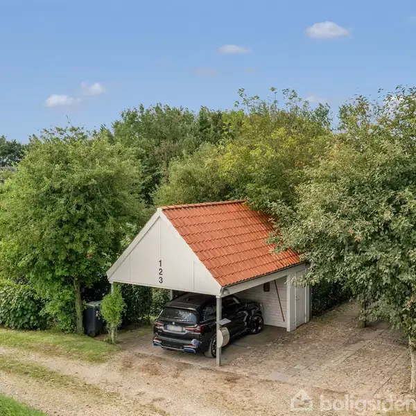 Carport med en sort bil parkeret under, omgivet af frodig beplantning og en grusvej. Taget har røde teglsten, og der står "123" på gavlen.