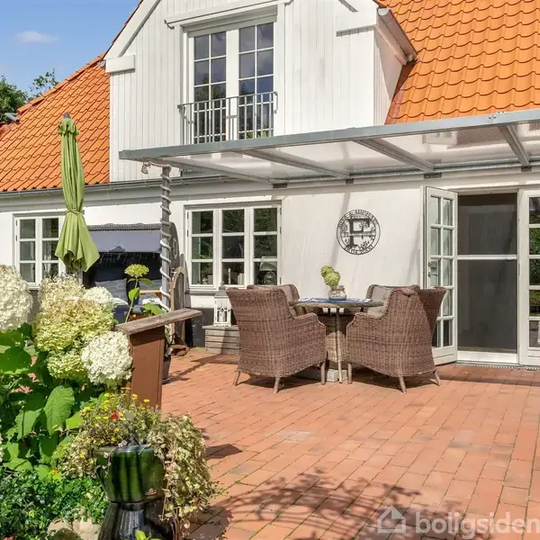 Et hyggeligt udendørs terrassemiljø med tremøbler og parasol, omgivet af blomstrende hortensiaer. Den tilhørende bygning har et rødt tegltag og store vinduer, der åbner til terrassen.
