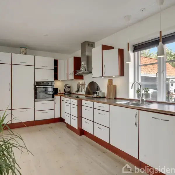 Et moderne køkken med hvide skabe og trækanter, hvor der står en ovn og komfur. Store vinduer giver udsigt til en have. En plante er placeret ved siden.