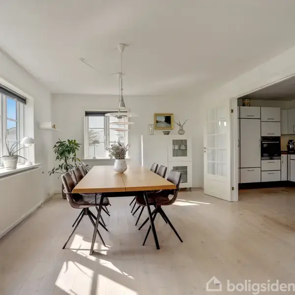 Et spisebord står midt i et lyst rum med seks stole omkring det. En vase står på bordet. Store vinduer og åbne glasdøre fører ind til et køkken.