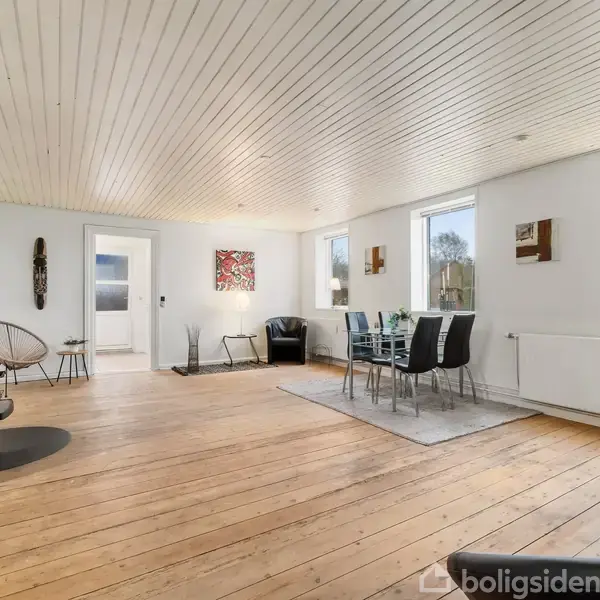 Stue med brændeovn til venstre, sofa og stol ved væggen, spisebord med stole på højre side. Lyse trægulve, hvidmalede vægge og stort vindue viser udendørs landskab.