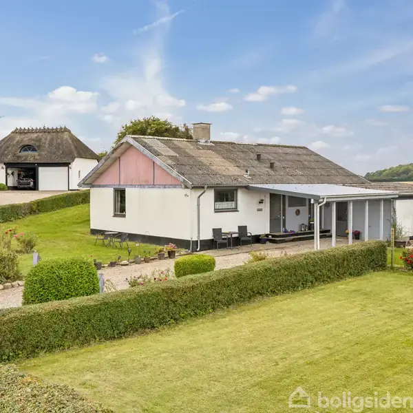 Et hvidt parcelhus med et skråt tag står i en velplejet have omgivet af hække. Ved siden af huset er en carport, og længere væk ses en traditionel bygning med stråtag.