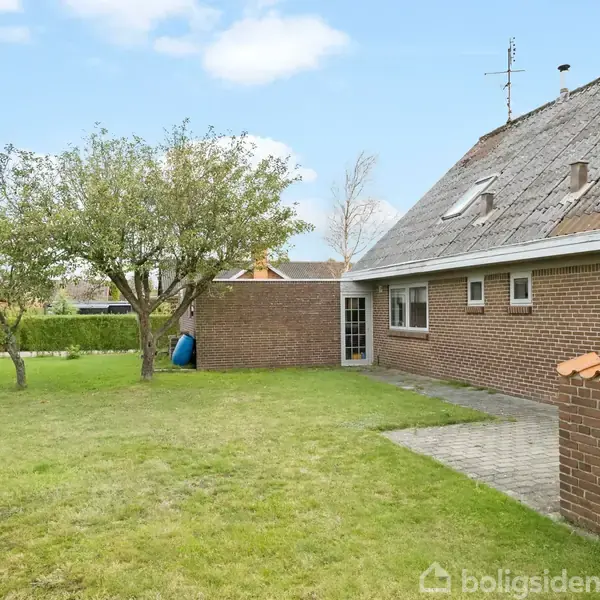 Hus i mursten med skråtag ses fra en have, hvor der står et træ. Et flisebelagt område fører til en dør ind til huset. Himmel er delvist skyet.