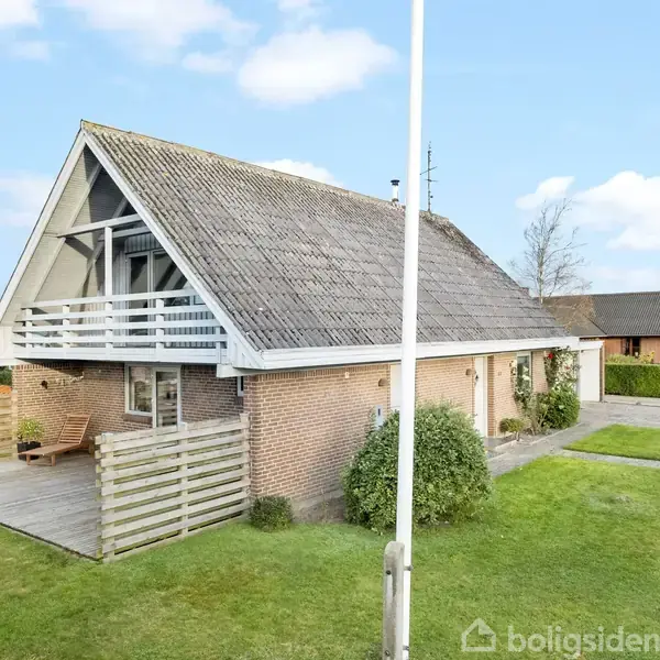 Et rødt murstenshus med en stor altan øverst og et fladt tagterrasseområde nederst. Haven er velplejet, med græs og buske, og omgivelserne inkluderer en vej og andre huse.