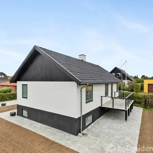Et moderne parcelhus med sort tag og hvide vægge, står på en flisebelagt grund. En lille veranda med glasrækværk strækker sig langs siden. Bakker og huse omkranser huset.