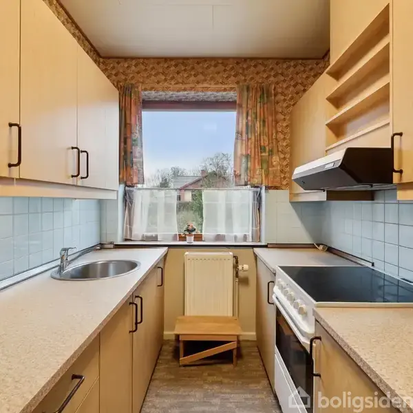 Køkkenet har lyse træskabe, en integreret vask, og en ovn. Et vindue med blomstrede gardiner leder ud til en have. Blå fliser dækker væggen over bordpladen.